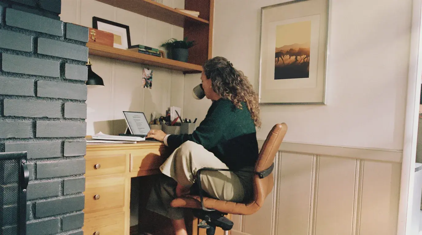 A person in a green jumper working at a laptop in a home office, a mug in one hand.